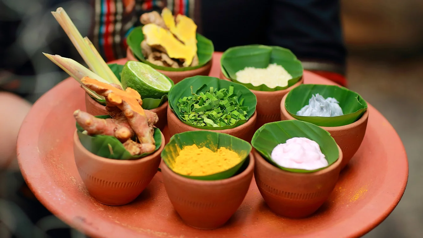 Thai herbal spa tray with turmeric, galangal, lemongrass, kaffir lime, and other natural ingredients, arranged in clay cups.
