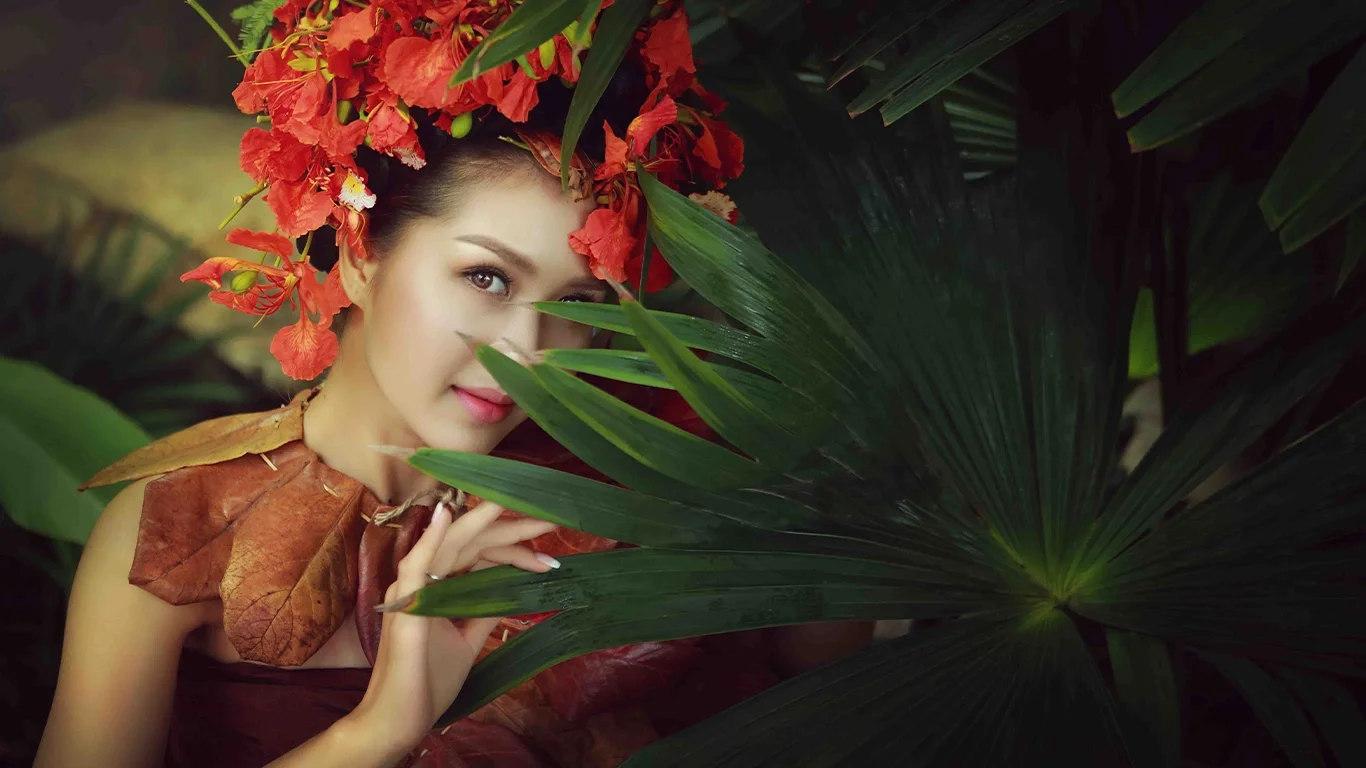 Woman in Lanna tribal attire adorned with red peacock flowers and dried leaves, gazing through green palm leaves, symbolizing natural beauty.