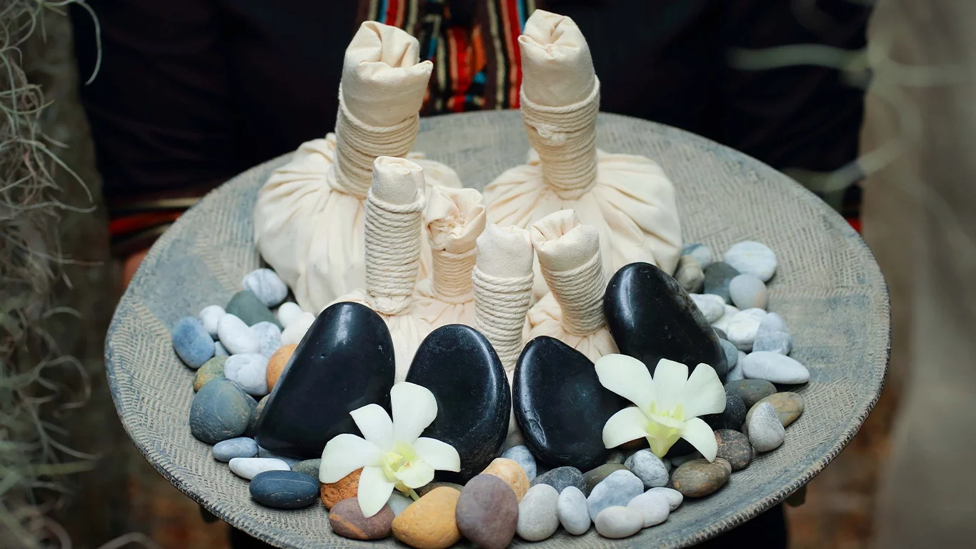 Herbal compress balls and spa massage stones arranged on a tray, adorned with white orchids, perfect for a relaxing massage.