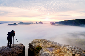 Professional nature photographer over clouds. Man takes photos with camera on tripod on rocky peak.